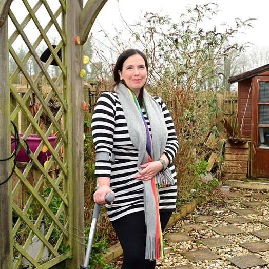 Councillor Laura Murtagh stands by a pergola with her walking stick in her hand and autumnal plants and her garden shed behind her