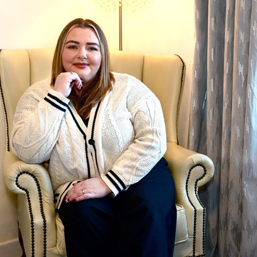 Councillor Stacey Devine sits in a white armchair looking directly towards the camera with a window to her right and a floor lamp with cream shade behind her